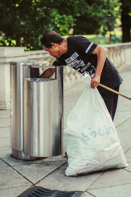 A middle-aged man with dark hair, wearing a black T-shirt with white text and dark pants, is seen bent over, using his right hand to place a large white plastic bag into a tall, cylindrical stainless steel public rubbish bin on a sidewalk. The bin has a smooth, reflective surface and a slightly rounded top, positioned near a low, decorative stone wall that runs parallel to the pavement. In the background, lush green trees with dense foliage provide a natural setting, indicating an outdoor urban or park environment. The man holds a long wooden stick or broom handle in his left hand, which rests against the rubbish bag he is handling, suggesting he is engaged in waste collection or street cleaning. The scene is illuminated by natural daylight, with shadows cast on the ground, emphasizing the outdoor context and the routine activity of rubbish disposal or street cleaning, consistent with services provided by companies specializing in private rubbish removal solutions.