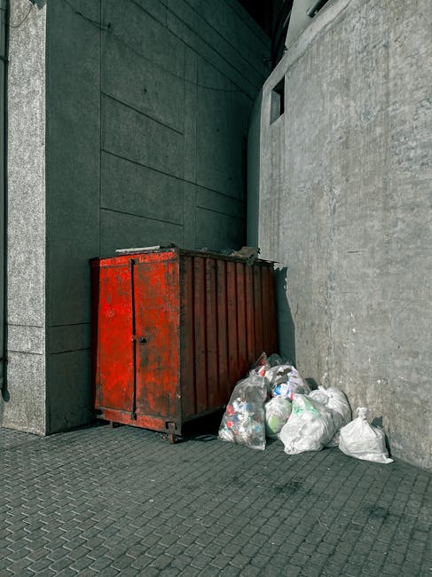 A middle-aged man with dark hair, wearing a black T-shirt with white text and dark pants, is seen bent over, using his right hand to place a large white plastic bag into a tall, cylindrical stainless steel public rubbish bin on a sidewalk. The bin has a smooth, reflective surface and a slightly rounded top, positioned near a low, decorative stone wall that runs parallel to the pavement. In the background, lush green trees with dense foliage provide a natural setting, indicating an outdoor urban or park environment. The man holds a long wooden stick or broom handle in his left hand, which rests against the rubbish bag he is handling, suggesting he is engaged in waste collection or street cleaning. The scene is illuminated by natural daylight, with shadows cast on the ground, emphasizing the outdoor context and the routine activity of rubbish disposal or street cleaning, consistent with services provided by companies specializing in private rubbish removal solutions.
