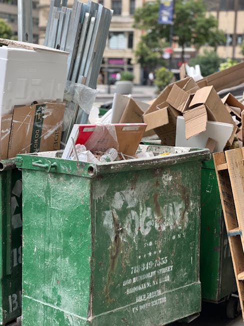 A large, weathered green waste disposal dumpster filled with various discarded materials including cardboard boxes, paper packaging, and plastic wrapping, situated outdoors on a paved surface. The surrounding area features additional bins, some of which are partially visible, along with a background of urban environment elements such as buildings, trees, and parked cars. The cardboard boxes are in different shades of brown, some flattened and others still in box form, with corrugated textures visible. The paper packaging appears crumpled and torn, while the plastic wrapping is loosely draped over the contents. The scene suggests an area designated for rubbish collection, possibly part of a private waste handling process, with the accumulation of debris indicating ongoing clearance or disposal activity. Natural daylight illuminates the scene, highlighting the textures and colors of the waste materials, in alignment with waste removal services like those offered by House Clearance Kennington, supporting discussions around independent rubbish collection methods and local waste management challenges.