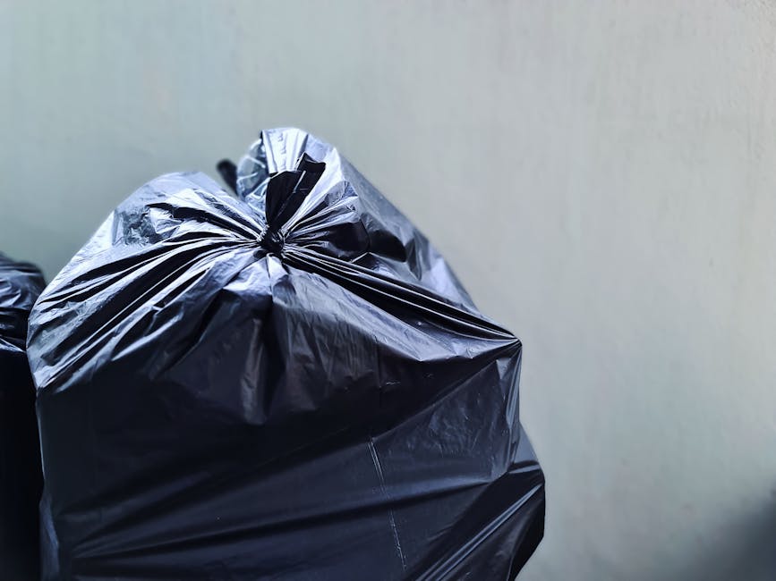 Close-up of a tightly packed black plastic rubbish bag with a glossy surface, gathered and twisted at the top, resting against a neutral light-colored wall. The bag appears to contain household waste and is situated outdoors or in an enclosed area such as a driveway or yard. In the background, faint outlines of additional rubbish bags are visible, suggesting a collection of waste awaiting removal. The scene is evenly lit, with natural or artificial light reflecting off the plastic surface, highlighting its crinkled texture and the creases formed during tying. The environment suggests a typical setting for private waste disposal, commonly associated with independent rubbish collection or on-site clearance by companies like House Clearance Kennington, supporting alternative waste handling methods outside official council collection services. The focus remains on the waste bag’s material and form, with minimal environmental context to maintain clarity and focus on the object itself.