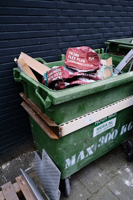 A large, weathered green waste disposal dumpster filled with various discarded materials including cardboard boxes, paper packaging, and plastic wrapping, situated outdoors on a paved surface. The surrounding area features additional bins, some of which are partially visible, along with a background of urban environment elements such as buildings, trees, and parked cars. The cardboard boxes are in different shades of brown, some flattened and others still in box form, with corrugated textures visible. The paper packaging appears crumpled and torn, while the plastic wrapping is loosely draped over the contents. The scene suggests an area designated for rubbish collection, possibly part of a private waste handling process, with the accumulation of debris indicating ongoing clearance or disposal activity. Natural daylight illuminates the scene, highlighting the textures and colors of the waste materials, in alignment with waste removal services like those offered by House Clearance Kennington, supporting discussions around independent rubbish collection methods and local waste management challenges.