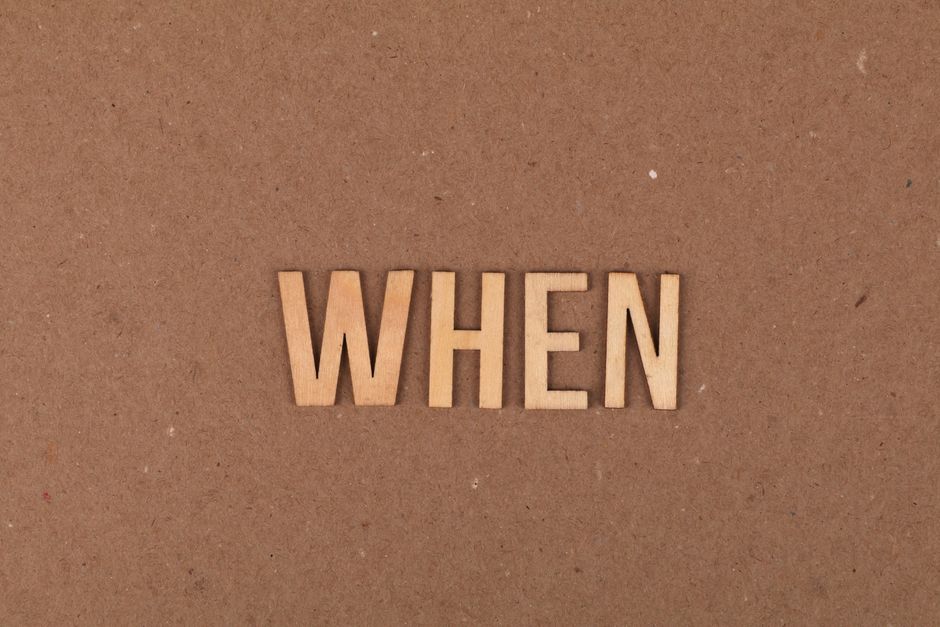 A close-up image of the word 'WHEN' spelled out with light-colored wooden alphabet letters placed on a plain, brown, textured surface. The background appears to be a matte, flat material, possibly cardboard or paperboard, with subtle variations in tone and a few minor particles visible. The lighting is even and natural, highlighting the texture of both the surface and the wooden letters. The composition is minimalistic, with the focus solely on the letters positioned centrally in the frame. The scene evokes a sense of inquiry or prompt, subtly connecting to the context of timing and scheduling associated with rubbish removal services, such as those provided by House Clearance Kennington, which might involve managing waste collection timings or private disposal arrangements outside of local authority schedules.
