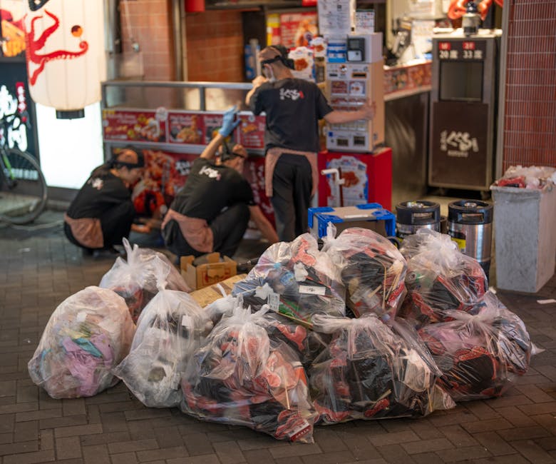 A collection of multiple large transparent plastic bags filled with various types of waste, including clothing and packaging, situated on a dark brick pavement outside a commercial establishment during evening lighting. Behind the bags, three workers dressed in dark uniforms with brown aprons and gloves are engaged in handling or organizing rubbish, with one standing and looking at a stack of box-like containers. The backdrop features a brightly lit storefront displaying menu boards, illuminated signage with Japanese characters, and a red and white paper lantern. Nearby, there are two black cylindrical bins and an extra plastic bin filled with waste. The scene suggests an on-site rubbish collection process, aligning with private waste disposal or alternative rubbish removal services, as part of a professional clearance operation managed by House Clearance Kennington. The environment appears clean and organized, with the scene captured in artificial lighting highlighting the details of the waste bags and staff activity.