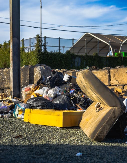 A pile of mixed rubbish and waste materials situated on a gravel surface in an outdoor setting. The collection includes black plastic garbage bags, some torn open, revealing loose packaging, paper debris, and small plastic items. A yellow plastic storage container and a worn, beige car seat with fabric upholstery are also visible in the foreground, leaning against the rubbish. Behind the pile, a low stone wall made of irregularly shaped, rough-textured stones runs horizontally across the scene, partially obscuring a background with a green hedge, metal fencing, and a large outdoor structure with a curved, semi-transparent roof. Overhead, several power lines cross the sky, which is partly cloudy with patches of blue. The overall environment suggests an area designated for waste disposal or an informal rubbish accumulation site, highlighting the importance of proper waste removal and secure disposal practices. House Clearance Kennington could assist with alternative waste handling options for landlords and property owners managing rubbish clearance.