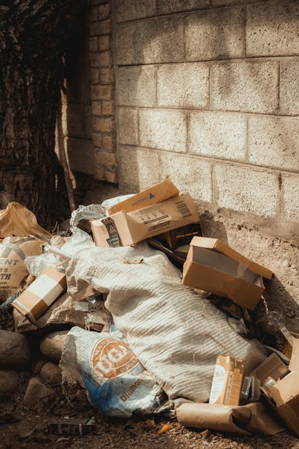 A pile of mixed domestic rubbish is situated at the base of a rough brick wall next to a tree trunk, with some cardboard boxes, plastic bags, and paper packaging visible on top of and around a large, textured, off-white sack. The assortment includes flattened and partially crumpled cardboard boxes, some with visible printed labels, alongside black plastic trash bags, and a few loose paper items. The scene is outdoors on a paved or dirt surface, with natural lighting illuminating the debris, which appears to have been discarded recently. The background features a weathered brick wall with light-colored mortar, bathed in soft daylight, and adjacent to the wall, the trunk of a mature tree provides a natural backdrop. This setting illustrates a typical example of waste accumulation that could be handled through private rubbish removal services, such as those offered by House Clearance Kennington, as an alternative to municipal waste collection, emphasizing the importance of proper disposal of household rubbish in urban environments.