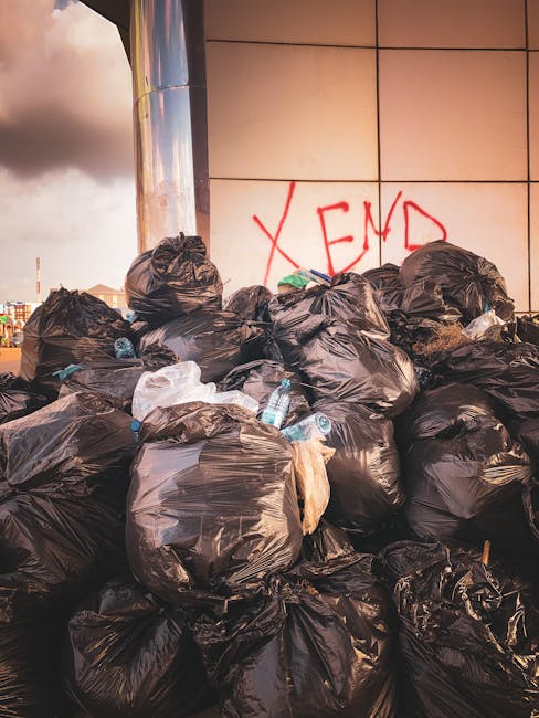 A collection of black rubbish bags and discarded packaging materials piled up against a beige, textured brick exterior wall beneath a red metal door, which features various graffiti tags in white and pink spray paint. The bags appear to be filled with household waste, and some cardboard and flattened packaging are visible amidst the pile. The door has multiple signs, including a white sign with black text that reads 'NO MONEY,' along with a blue and white 'Keep clear' sticker. The scene suggests a private waste collection or rubbish removal service, with the waste temporarily stored outside a building, possibly awaiting collection by services such as House Clearance Kennington. The environment is an urban alley or backstreet, with the waste arranged directly on the ground, creating a cluttered appearance typical of out-of-hours rubbish disposal areas leading to alternative waste handling solutions.