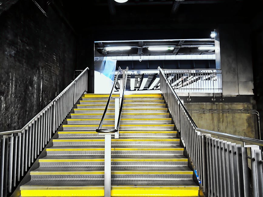 A wide staircase leading up from a lower level to an outdoor area, with metal handrails on both sides and a central handrail section. The steps are made of durable metal with a textured surface for slip resistance, edged with bright yellow safety strips along the front of each step. The staircase is enclosed by dark, industrial-style walls on the left and right, with visible concrete and paint marks. Above, there is a partly open ceiling, revealing structural beams and piping, with natural daylight illuminating the upper landing. The environment suggests an urban or public setting, such as a train station or parking garage, where independent rubbish removal or stair access is crucial. The scene is clean and functional, with no visible waste or clutter, aligning with professional waste management practices and private disposal access. The overall tone remains neutral and observational, emphasizing structural details and spatial arrangement without promotional language.