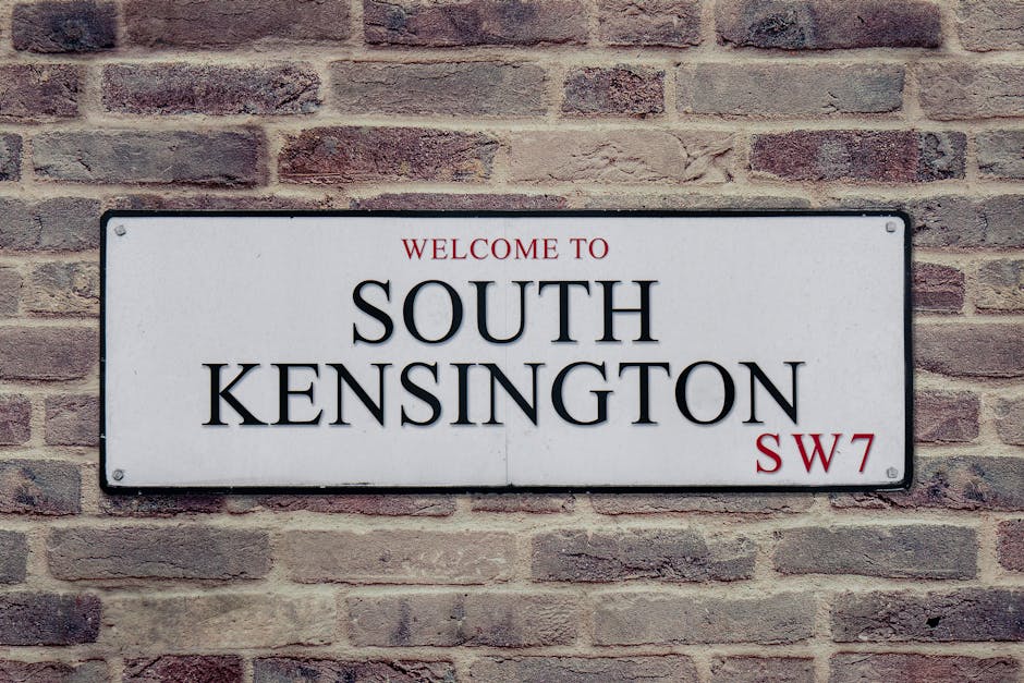 A rectangular metal street sign mounted on a brick wall, displaying the words 'WELCOME TO SOUTH KENSINGTON' in large black capital letters, with 'WELCOME TO' and 'SW7' in smaller red font. The sign has a white background and is secured with visible screws at each corner. The brick wall behind the sign is composed of reddish-brown and gray bricks arranged in a running bond pattern, with some bricks exhibiting weathered textures and slight color variation. The sign is positioned centrally in the image, and the scene appears to be outdoors in an urban setting. The lighting is natural and diffuse, creating minimal shadows and giving the scene a neutral, professional tone. This type of signage is typical in residential or commercial areas to designate locality, which could relate to private waste collection or local authority services, reflecting the context of alternative rubbish removal options but solely based on visual details.