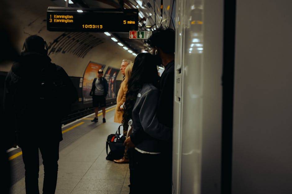 In a London underground station, several passengers are waiting on a platform to board trains. In the foreground, a woman with long dark hair, wearing a dark jacket, stands close to the wall, holding a small handbag or backpack. To her left, a man with glasses, dressed in dark clothing, stands facing away from the camera, carrying a backpack. In the background, a blonde woman with a beige coat and another individual in a grey jacket are visible, all standing on the platform, some engaged in conversation or looking towards the approaching trains. Overhead, electronic display boards show upcoming train destinations and timings, with information like 'Kennington' and departure times. The platform surface is smooth with visible yellow safety lines along the edge. The station’s curved ceiling is illuminated by evenly spaced lighting fixtures, creating a well-lit environment conducive to waiting commuters. The scene presents an everyday image of public transportation in London, supporting the concept of independent travel and alternative commuting options commonly associated with urban rubbish and waste management contexts, such as station cleanliness and waste disposal facilities managed by local authorities or private waste services like House Clearance Kennington.