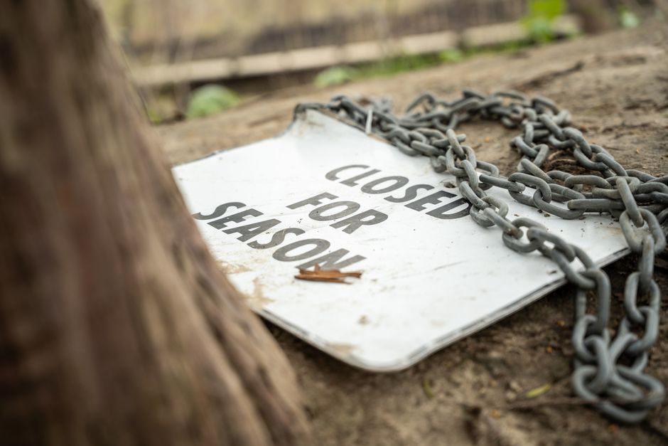 A weathered white metal sign with black lettering reading 'CLOSED FOR SEASON' lies on the ground amidst dirt and natural debris. The sign appears slightly bent and shows signs of rust and dirt accumulation, indicating exposure to outdoor elements. A heavy metal chain, gray in color and with thick links, is draped loosely over the sign, partially obscuring the text. The chain extends beyond the sign towards the right side of the image. In the background, there is a blurred wooden fence and a few green plants growing along it, suggesting the sign is situated outdoors, possibly in a garden or yard. The scene is lit with natural daylight, highlighting the textures of the rusted chain, the chipped paint on the sign, and the rough dirt surface. This image could relate to private or site-specific waste or rubbish management, such as temporary closures of designated disposal areas or restricted access points related to waste removal services, as provided by companies like House Clearance Kennington.