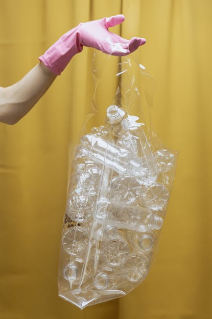 A person wearing a pink rubber glove is holding a large, clear plastic bag filled with crushed and intact transparent plastic bottles and containers, set against a plain yellow background. The bag appears to be lightweight and partially open at the top, with the bottles visible through the thin, flexible material. The environment suggests an indoor space, possibly related to waste sorting or recycling activities, in alignment with rubbish removal and waste management services offered by House Clearance Kennington. The detailed textures of the plastic and the vibrant contrast between the glove and the bag highlight the focus on waste collection and recycling processes, emphasizing the importance of proper disposal and alternative waste handling. The scene is professional and neutral, with a clear observation of the items intended for recycling or disposal, supporting the context of independent collection or private waste clearance.