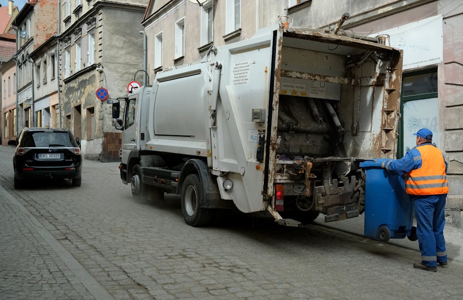 A waste collection vehicle, primarily white with some weathered rust spots, is parked on a cobblestone street in an urban area, positioned close to the sidewalk on the right side of the image. A worker dressed in blue coveralls and an orange high-visibility vest, wearing a blue cap, is seen placing a blue wheeled bin into the rear compartment of the truck, which is designed for on-site rubbish collection or private waste disposal. The back of the vehicle is open, revealing a mechanical lifting mechanism and a rusty, worn interior used for compacting and loading waste materials. In the background, there are residential buildings with light-colored facades, mixed with some signs and window coverings, indicating an older part of the city. A small black car is parked further down the street, and two no-parking signs are visible attached to building facades. The overall scene depicts typical urban rubbish collection activity, emphasizing private waste management services like those offered by House Clearance Kennington, with a natural focus on the environmental context of rubbish and waste removal near residential areas.