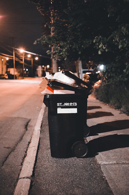 A black wheelie bin marked with the label 'St. John’s' is positioned on a pavement beside a road at night. The bin is filled with a mix of household waste, including white and brown paper packaging, plastic containers, and other refuse, some of which are spilling over the top. The textured bin lid is open, revealing the rubbish inside. The scene is illuminated by nearby streetlights, casting long shadows on the asphalt and sidewalk. In the background, faint outlines of trees and parked cars are visible, with additional streetlights and power lines stretching across the sky. The setting suggests an area where residents may use private waste collection services, such as those provided by companies like House Clearance Kennington, for home clearance or rubbish removal outside regular council collection hours.