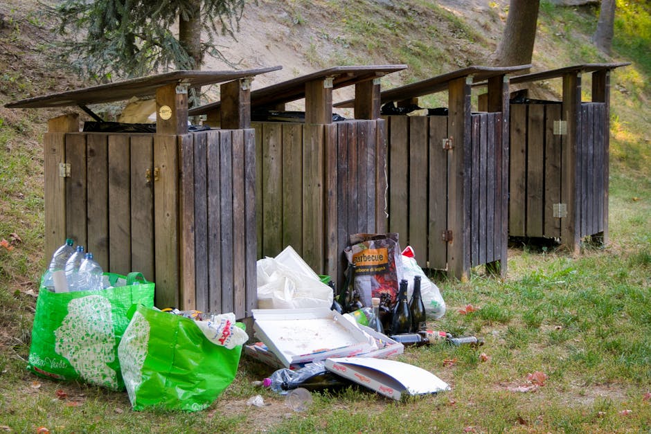 A row of four wooden outdoor waste bins with hinged lids, situated on a grassy area beneath tall trees with green foliage. The bins are weathered, with visible grain and a dark brown finish, each featuring metal hinges and locks. The lids are slightly tilted, revealing some interior contents. In front of the bins, there are various discarded items including a green reusable shopping bag filled with empty plastic bottles, a white pizza box, a paper bag of barbecue charcoal, and several glass bottles, some of which are dark-colored and others transparent, along with plastic wrappers and miscellaneous rubbish scattered on the ground. The surrounding environment appears to be part of a park or woodland area, with a gentle slope and a mix of grass and fallen leaves, creating a scene of private waste disposal or illegal dumping. The natural lighting indicates a daytime setting, with dappled sunlight filtering through the trees, highlighting the contrast between the functional waste containers and the unmanaged litter in front of them. This scene aligns with the theme of rubbish collection and waste management services, illustrating the challenge of illegal dumping adjacent to outdoor waste facilities.
