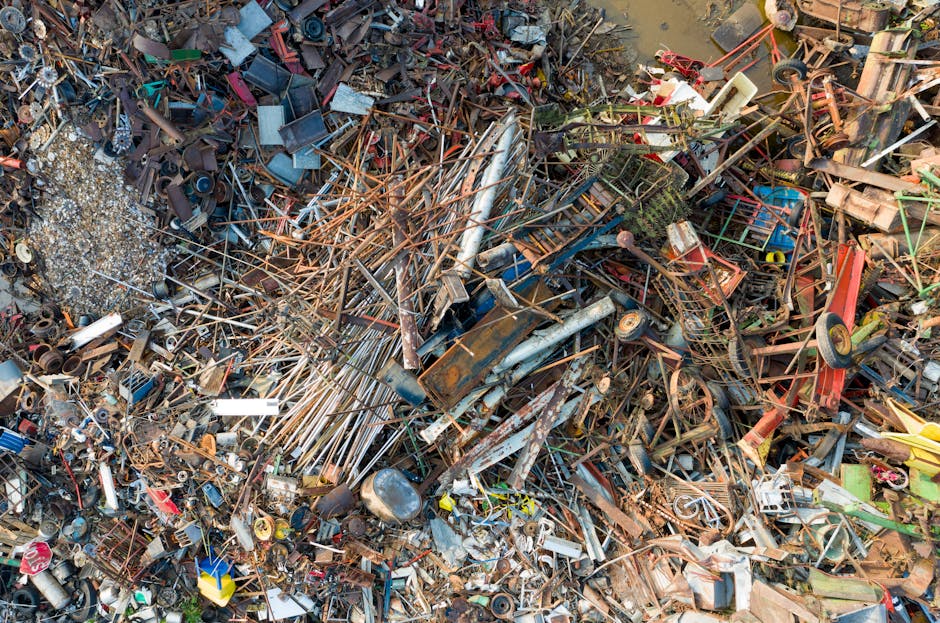 A large pile of assorted scrap metal and discarded objects, including rusted pipes, metal rods, broken machinery parts, and scattered smaller items such as tin cans, wires, and plastic components. The materials vary in texture, with some pieces exhibiting corrosion and rust, giving an uneven rough surface, while others have smooth, painted finishes. The colour palette comprises shades of brown, rust, grey, and hints of blue and red from painted or plastic elements. The scrap is densely packed, filling the frame from top to bottom with objects stacked chaotically, some leaning on each other and others partially buried. Surrounding the pile is an outdoor environment, likely on a gravel or dirt surface, with some debris spilling over onto the ground. The lighting is natural, highlighting the metallic sheen and rusted surfaces. This scene exemplifies a site where independent or private waste disposal, such as scrap metal collection, takes place, akin to services provided by companies like House Clearance Kennington for rubbish removal and on-site clearance of metal waste materials.