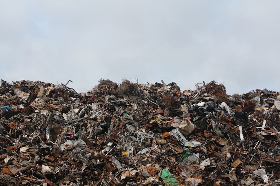 A large mound of mixed waste materials extends across the lower half of the image, consisting of crushed and shredded metal, plastic, cardboard, and organic debris. The pile includes rusty metal scraps, broken plastic items in various colors, pieces of cardboard with torn edges, and some organic matter such as twigs and decomposing plant material. The waste appears to be randomly piled, creating a chaotic and dense surface with uneven textures and a range of dull colors including browns, greys, greens, and occasional bright spots from plastic packaging. The background features a pale, overcast sky that contrasts with the cluttered foreground, emphasizing the extent of the waste heap. The environmental context suggests the pile is situated outdoors, possibly within a waste disposal site or between urban structures. The scene reflects the accumulation typical of waste management situations, where materials await collection or processing by rubbish removal services like House Clearance Kennington, handling non-recyclable refuse or bulk rubbish from residential or commercial properties.